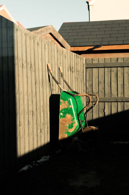 A green wheelbarrow with a rusty metal handle resting against a tall wooden fence in a backyard garden. The fence is made of vertical natural wood planks, and behind it, parts of a neighbouring house 